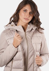 Giacca imbottita beige chiaro con zip frontale, cappuccio regolabile e polsini elasticizzati. Presenta sezioni trapuntate e dettagli in metallo color oro.