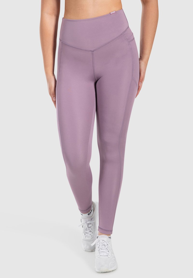 Legginsy z wysokim stanem w jasnym fioletowym kolorze, wykonane z elastycznego materiału, z szerokim pasem w talii i detalami przeszycia po bokach. W połączeniu z białymi trampkami.