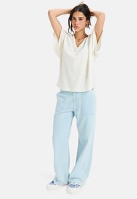 Camisa blanca de pico con mangas anchas, combinada con pantalones de pierna ancha de color azul claro. La modelo lleva zapatillas blancas con acentos azules.
