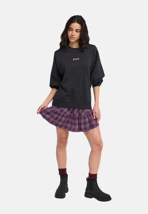 Vrouw die een zwart shirt met lange mouwen draagt met de tekst "ROXY", een korte rode geruite rok, bordeauxrode sokken en zwarte enkellaarzen.