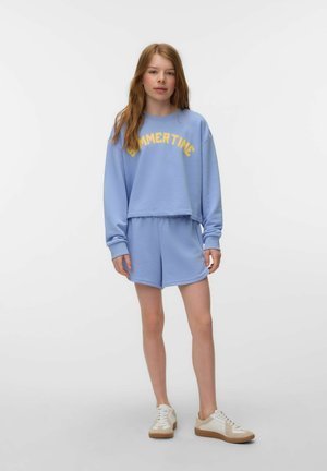 Girl standing wearing light blue sweatshirt with "SUMMERTIME" text and matching shorts with white sneakers on a white background.
