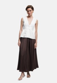 Biała bezrękawnikowa bluzka peplum z głębokim dekoltem w kształcie litery V i detalami z guzików, w zestawieniu z długą brązową spódnicą. Modelka nosi lekkie sandały.