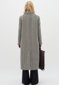 Dlhý kabát s houndstooth vzorom v čiernej a krémovej farbe, s klopovým golierom a širokými rukávmi, skombinovaný s prispôsobenými čiernymi nohavicami a tmavo hnedou taškou.