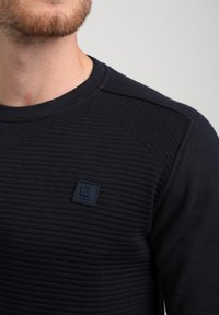 Felpa blu navy con texture a coste sulla parte anteriore, maniche lisce e un'etichetta a logo quadrato sulla parte sinistra del petto.