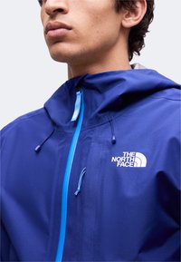 Jeune personne portant une veste à capuche The North Face bleue avec une fermeture éclair bleu clair et des cordons de capuche réglables.