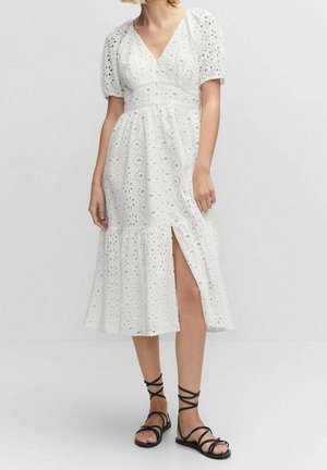 Kobieta ubrana w białą sukienkę midi z ażurowym wzorem, bufkami na rękawach i bocznym rozcięciem, w połączeniu z czarnymi płaskimi sandałami na sznurowadła.