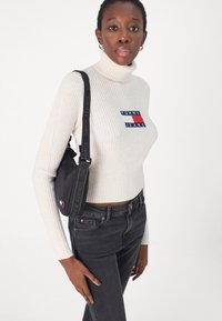 Maglione a coste color crema con collo alto e patch con logo, abbinato a jeans neri. Una piccola borsa nera con tracolla di logo pende dalla spalla.