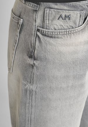 Detalle de unos jeans de mezclilla gris claro que muestra el bolsillo delantero con el logo bordado "AM" en el pequeño bolsillo para monedas y costuras visibles en la costura lateral.