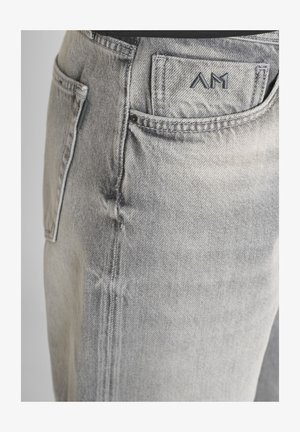 Svijetlosivi traperici s detaljem prikaza prednjeg džepa s ušivenim "AM" logom na malom džepu za novčiće i vidljivim šavovima na bočnoj strani.