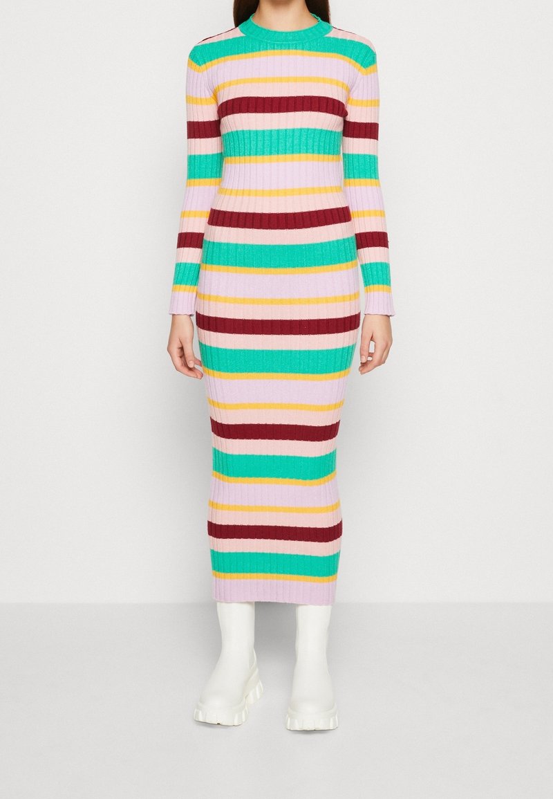 Femme portant une robe longue côtelée multicolore à rayures avec des manches longues et des bottes blanches épaisses, debout devant un fond uni.