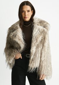 Woman wearing a spotted faux fur coat over a black turtleneck and black pants with a belt, posing with one hand in pocket.
