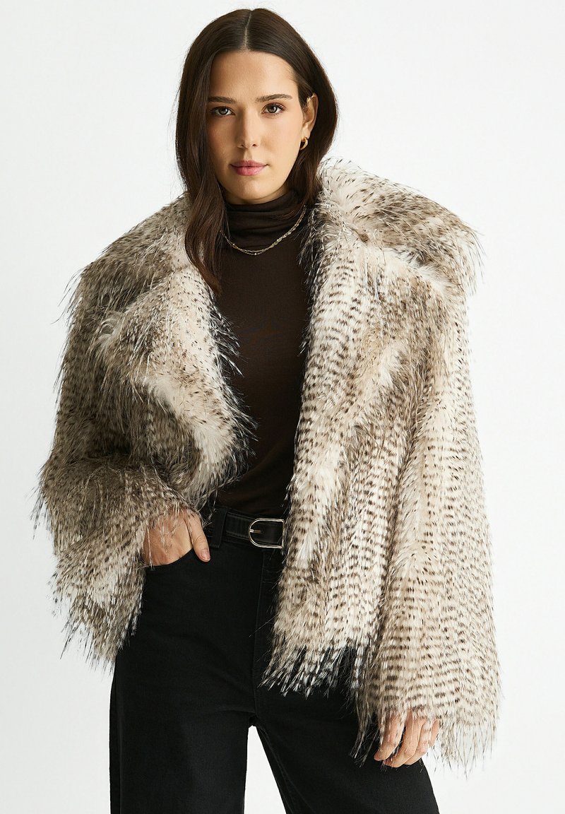 Woman wearing a spotted faux fur coat over a black turtleneck and black pants with a belt, posing with one hand in pocket.