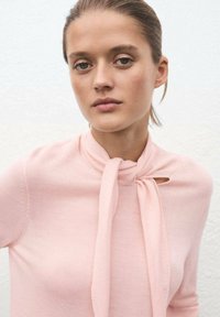 Maglione rosa chiaro a maglia con un fiocco al collo e un dettaglio a chiave. Texture liscia, design aderente e maniche lunghe.