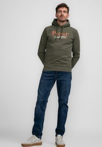 Hoodie verde oliva con testo del logo bianco e arancione, abbinato a jeans in denim blu. Presenta un cappuccio con cordini e una vestibilità comoda.