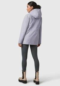 Giacca impermeabile di un leggero colore lavanda con cappuccio, tasche laterali e vita leggermente stretta. Indossata con jeans scuri e stivaletti beige.