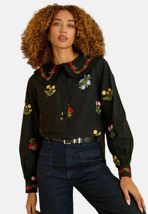 Black blouse with embroidered floral patterns in red, yellow, and blue, featuring a ruffled collar and button-down front, paired with dark jeans.