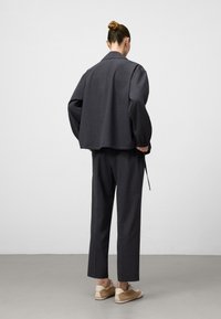 Persoon met een bun kapsel, gekleed in een donkergrijze losse jas en bijpassende broek, gecombineerd met beige en witte sneakers, staande op een grijze vloer.