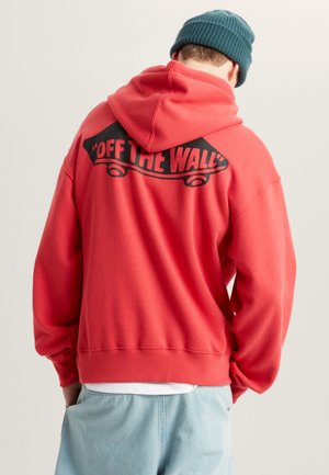 Felpa rossa con una grande grafica nera "OFF THE WALL" sul retro. Presenta un cappuccio con cordino e polsini a coste, indossata con pantaloni azzurri chiari.