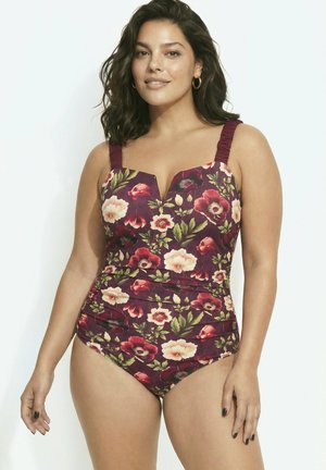 Frau mit mittellangen dunklen Haaren, die einen dunkelroten, geblümten Einteiler mit breiten gerafften Trägern trägt, steht vor einem weißen Hintergrund.