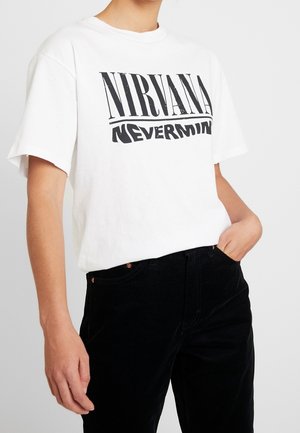 Osoba ubrana w białą koszulkę z czarnym tekstem "Nirvana Nevermind" i czarne spodnie, ręka w kieszeni na jednolitym tle.