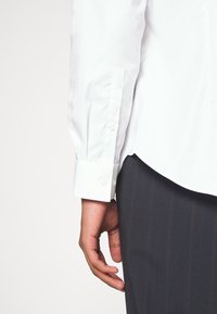 Chemise blanche à manches longues avec des poignets boutonnés, présentant une texture lisse. Associée à un pantalon rayé foncé, montrant le détail des poignets et la position des mains.