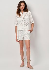 Witte katoenen blouse met oogjes, korte mouwen en twee voorzakken, gecombineerd met bijpassende shorts. De outfit heeft uitsnijdingen en een relaxte pasvorm.