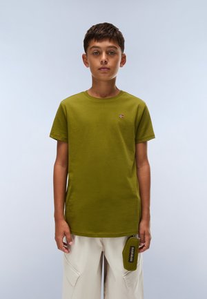 Un garçon porte un tee-shirt vert olive et un pantalon cargo couleur crème, en tenant une pochette verte assortie devant un fond uni bleu clair.