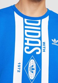 Camiseta azul de manga corta con rayas verticales blancas que lleva "ADIDAS" y un logo, confeccionada con una tela lisa y ligera.