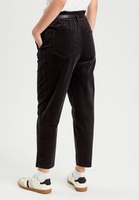 Pantalons en velours côtelé noirs avec une coupe décontractée, dotés d'une ceinture élastique, d'un design à cinq poches et de jambes fuselées, associés à des baskets blanches.