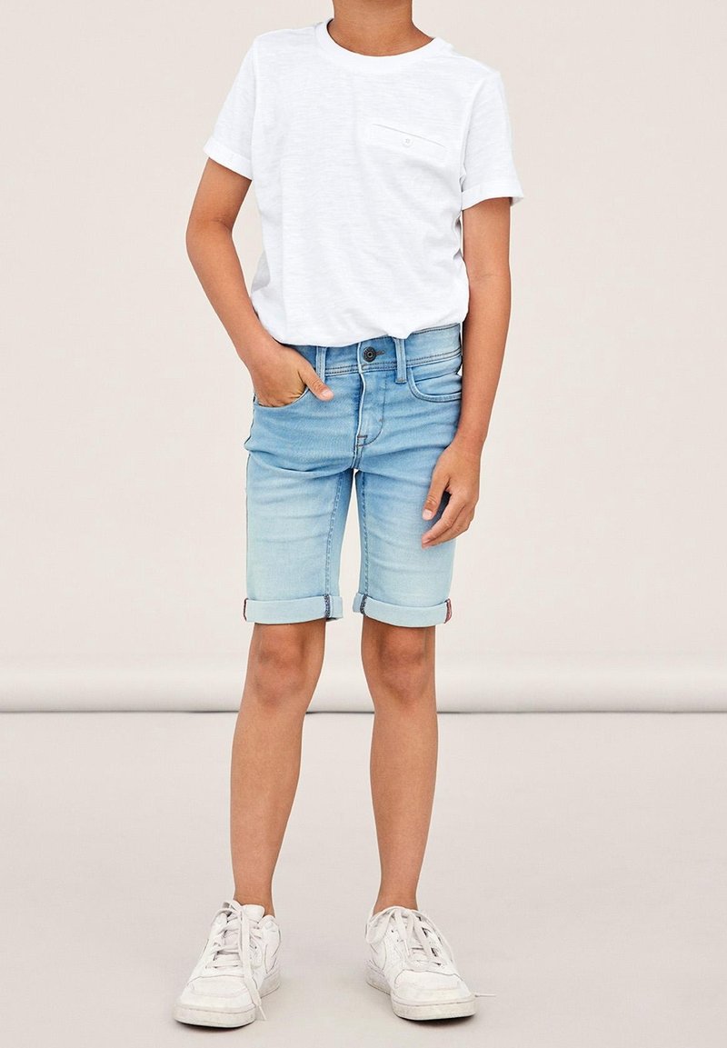 Enfant portant une chemise blanche à manches courtes, un short en denim bleu clair retroussé et des baskets blanches, debout avec une main dans la poche.