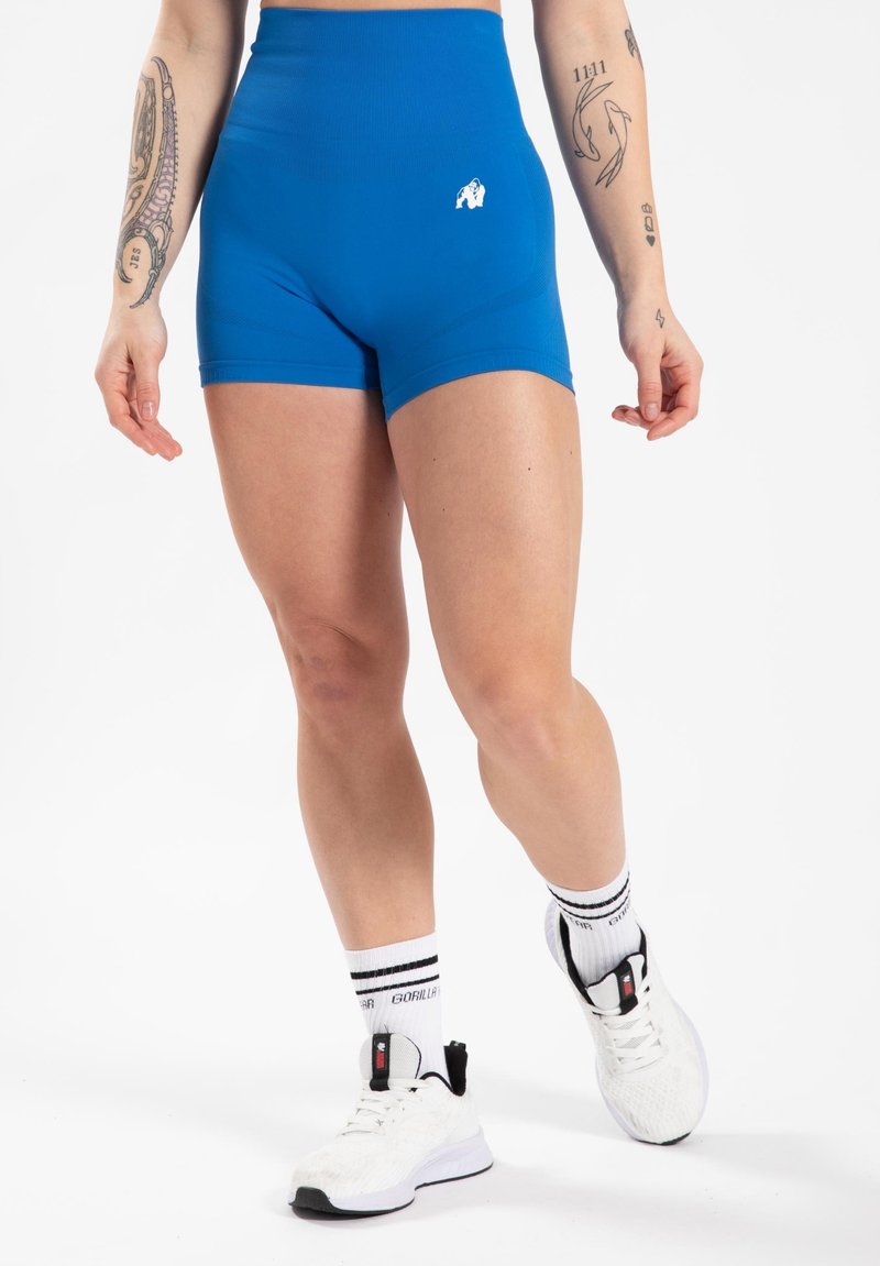 Blauwe hoge taille shorts van rekbaar materiaal, met een klein wit logo, gecombineerd met witte sneakers en gestreepte sokken.