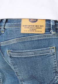 Blauwe denim jeans met een bruine leren label op de achterkant, voorzien van gele stiksels en een standaard zakontwerp. De stof heeft een lichte textuur.
