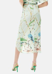 A mint green midi skirt with a botanical and bird pattern, featuring a side slit and an elastic waistband for comfort. Paired with green heels.