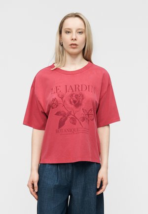 Kvinde med glat blondt hår iført en stor rød T-shirt med botanisk tryk af roser og sommerfugle samt løstsiddende mørkeblå jeans.