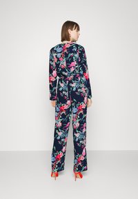 Jumpsuit floral com fundo azul-marinho e padrões vibrantes em rosa, azul e verde, com mangas longas e detalhe de cintura atada.