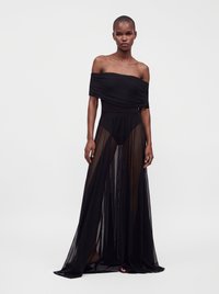 Femme debout portant une robe noire épaules dénudées avec une jupe ample en voile transparent par-dessus un short ajusté, sur fond gris clair uni.