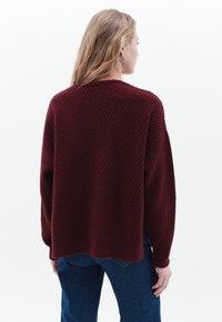 Bordeaux gebreide trui met een gestructureerd visgraatpatroon, verlaagde schouders en geribbelde manchetten, gecombineerd met blauwe jeans.