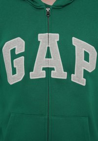 Πράσινο φούτερ με φερμουάρ, με γκρι λογότυπο "GAP" από τσόχα στο στήθος, μπροστινές τσέπες και ριμπ μανσέτες και τελείωμα. Το φερμουάρ είναι ασημί.