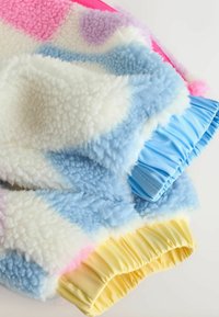 Krāsaina vilnas sega ar mīkstām tekstūrām rozā, zilā, baltā un dzeltenā krāsā, ar savāktām auduma malām. Akcenti ietver pasteļtoņus.