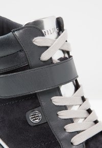 Close-up of a stylish black sneaker featuring suede material, gray accents, metallic details, and light-colored laces on a white background.