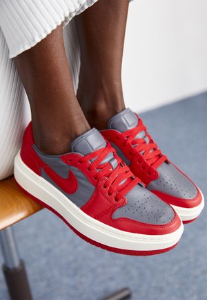 Pieds portant des baskets Nike grises et rouges avec des lacets rouges, reposant sur une chaise en bois, personne portant un pantalon blanc plissé.