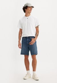 Weißes kurzärmeliges T-Shirt, blaue Jeansshorts, weiße Sneakers und eine graue Mütze. Tattoos sichtbar an den Armen, steht vor einem schlichten Hintergrund.