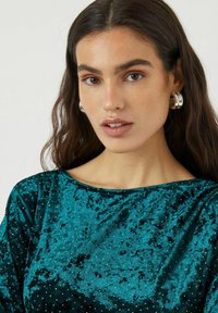 Tyrkysová sametová blůza s lodičkovým výstřihem, zdobená malými stříbrnými korálky na látce, vytvářejícími texturovaný vzor.
