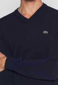 Hluboká modrá svetr s výstřihem do V, ribovanou texturou a dlouhými rukávy. Obsahuje malé zelené logo Lacoste na levé hrudi.