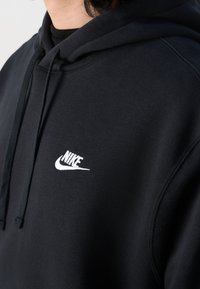 Černá mikina s kapucí vyrobená z měkké tkaniny, na hrudi má bílou značku Nike. Má stahovací kapuci a manžety s žebrováním.