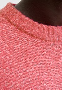 Close-up van een koraalroze gebreide trui met een halslijn op een zwarte mannequin, met zichtbare ribbelkraag en een gestructureerd breipatroon.