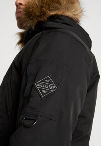 Černá parka s kapucí podšitou kožešinou, s diamantovým výšivkou s nápisem "HOLLISTER" a "1999" na levém rukávu. Obsahuje popruh a detail s kroužkem.