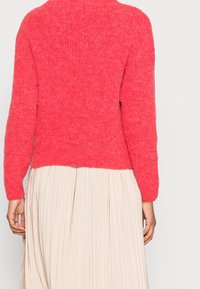 Luddig, korallfärgad pullovertröja med hög halsringning och långa ärmar, kombinerad med en plisserad, beige kjol. Mjuk textur med lös passform.