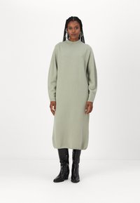 Robe tricotée verte clair à manches longues avec une coupe décontractée et des poignets à côtes, associée à des bottes noires brillantes jusqu'au genou.