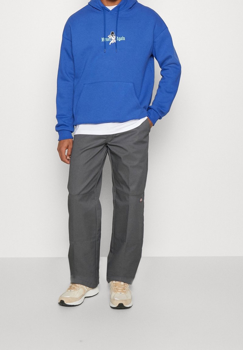 Felpa con cappuccio blu con logo, pantaloni grigi e sneakers chiare. La felpa ha una tasca a marsupio; i pantaloni hanno tasche laterali e una toppa con logo.
